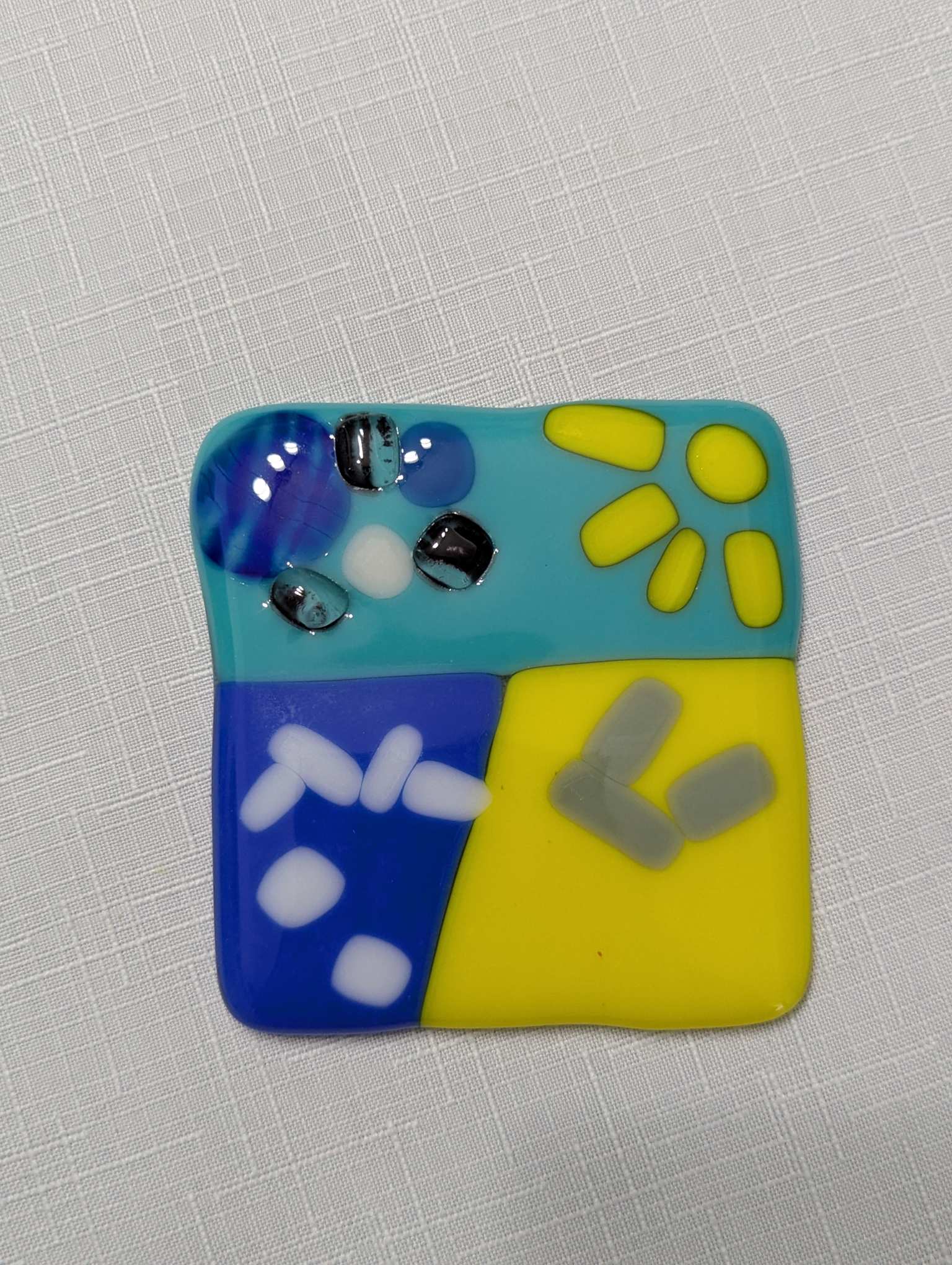 Fused glass landscape of a beach, with gentle waves on the water. The sun shines over the beach, with dark clouds looming over the water. Lying on the beach are robotic legs and a hip connection.