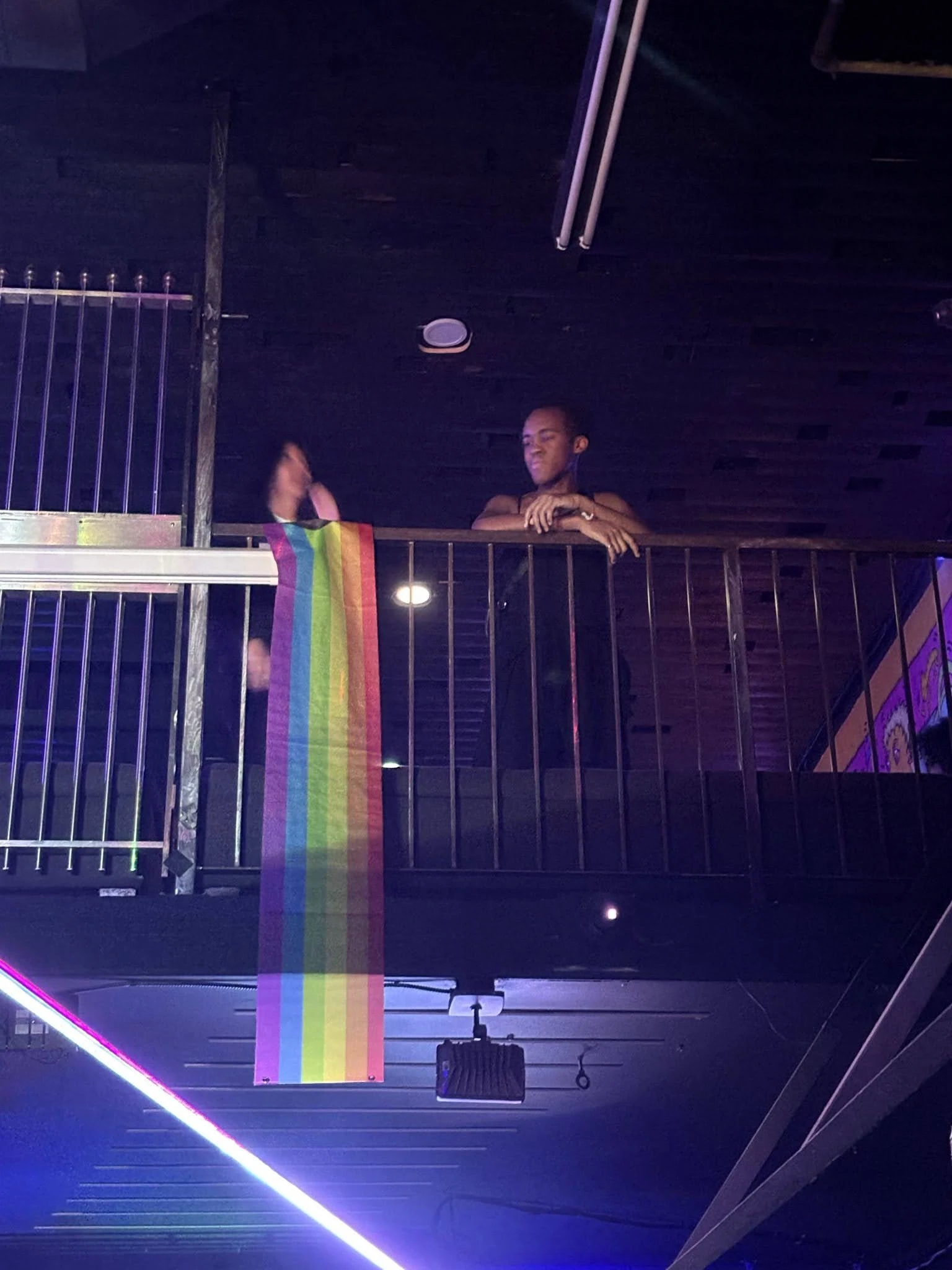 Jessica, wearing a black dress, leans over the upper balcony in a club. A laser light shoots from the lower floor, and a Pride flag is draped from the balcony railing.