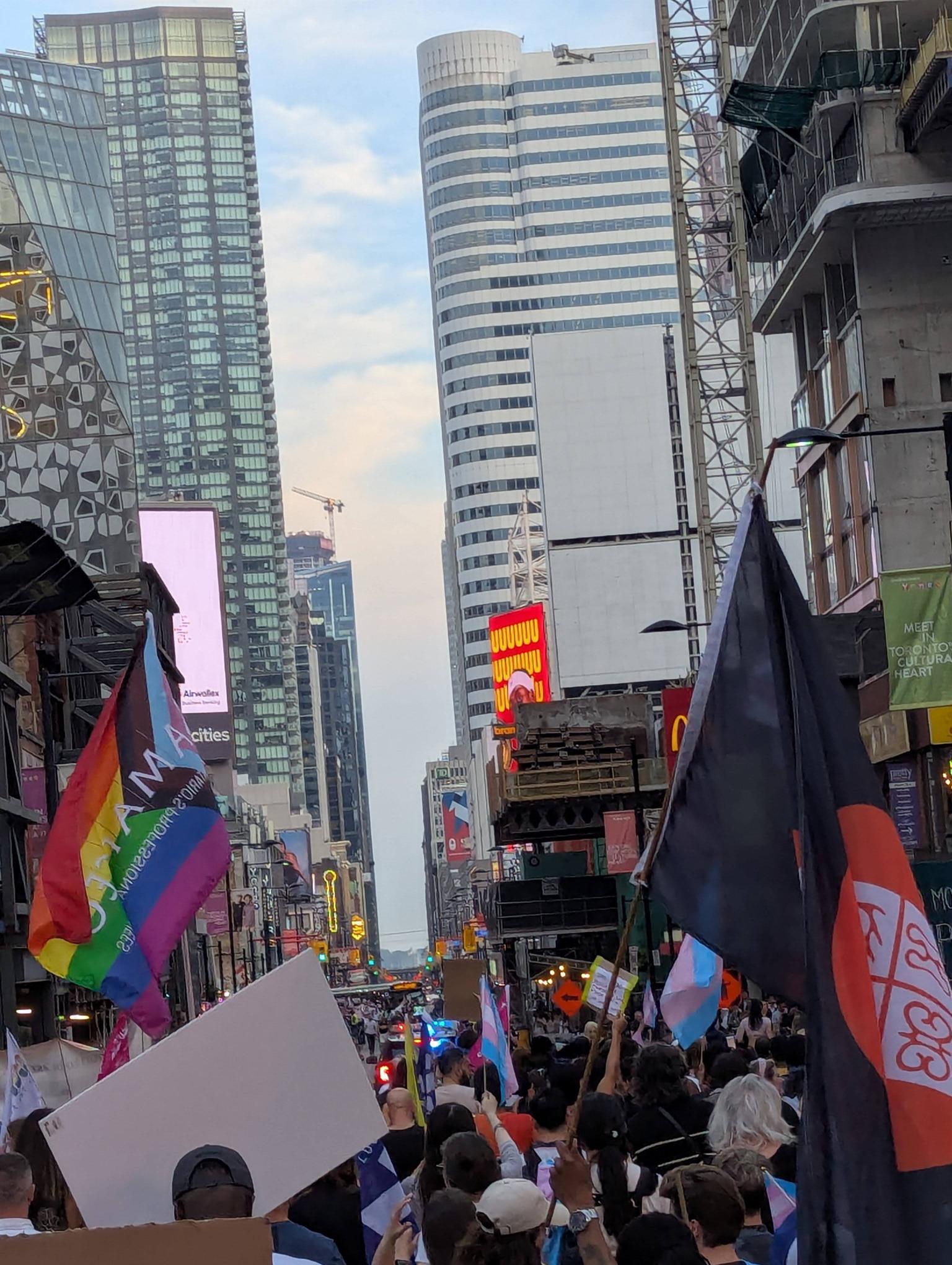 A photo taken facing down Yonge Street during the Toronto Trans March. Hundreds of people are marching or cheering, waving flags all the way down to Dundas Street.