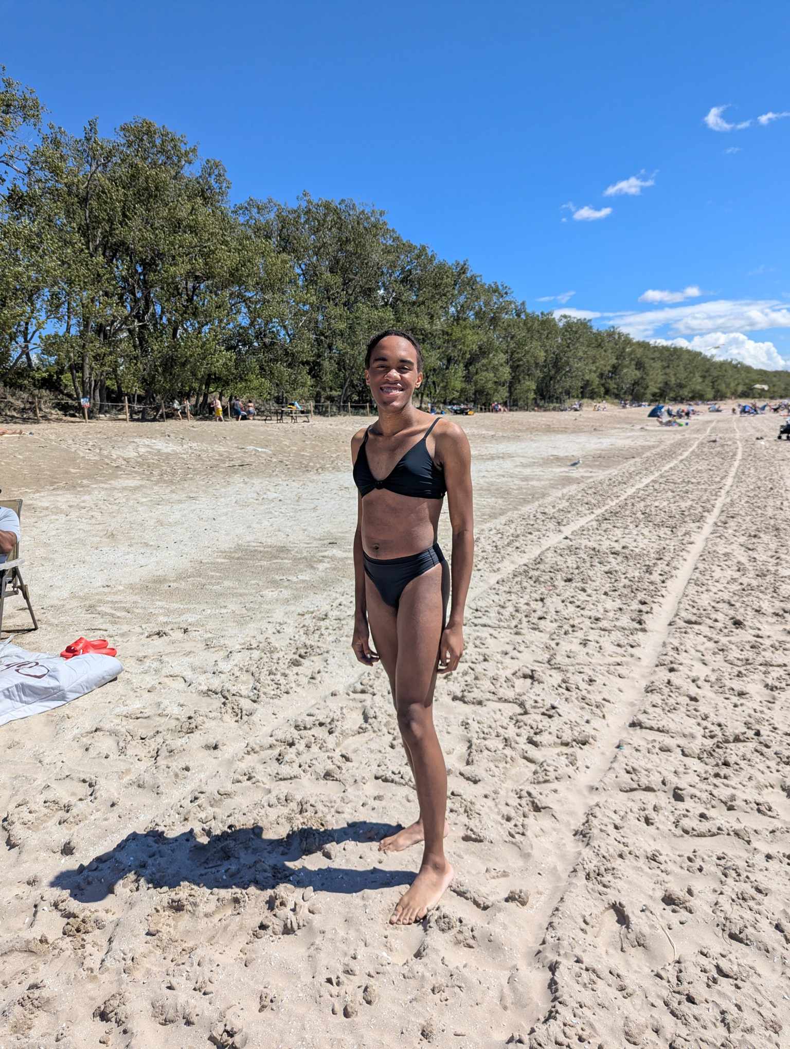 Jessica is wearing a black bikini, smiling at the camera, with her body angled slightly to the left. She's standing on the beach, with a treeline along the entrance in the background.